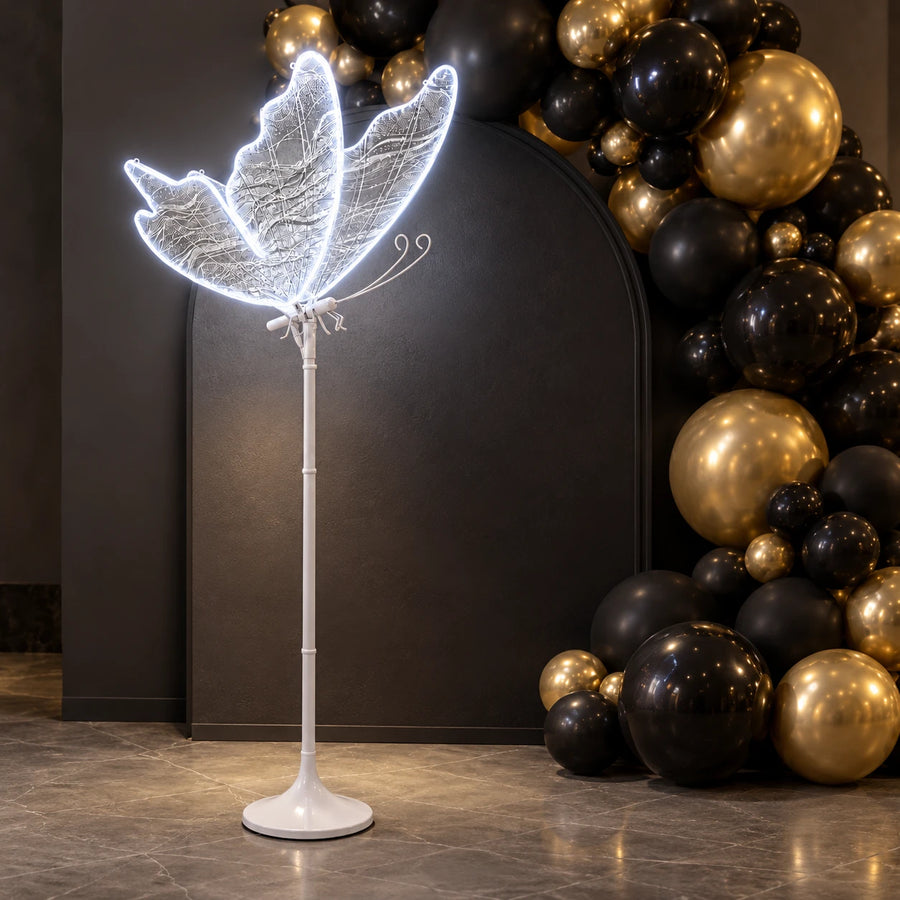 Lampa Motyl LED świeci zimną bielą przy ścianie z czarnymi i złotymi balonami
